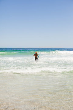 Rear view of a blonde man running in the water with his surfboar