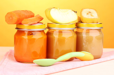 Jars of baby puree with spoon on napkin on yellow background