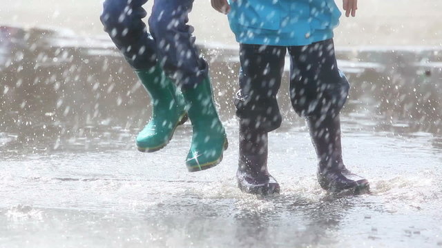 Unrecognizable children jumping in puddle during rain, wearing rubber boots, splashing water. Suitable for lifestyle, kids activities and seasonal content