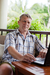 mature man sits on a balcony of tropical hotel with the laptop