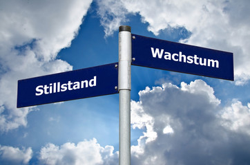 Straßenschild vor bewölktem Himmel welches den Zielkonflikt zwischen 'Stillstand' und 'Wachstum'...