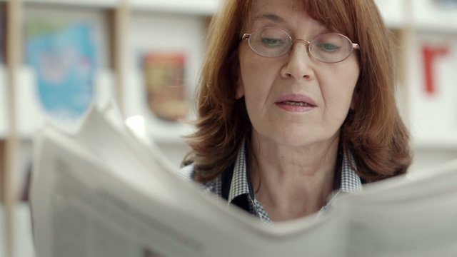 Old woman with glasses reading newspaper in library
