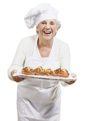 senior woman cook holding a tray with muffins against a white ba