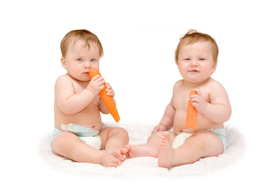 two lovely chubby baby twins sitting eating an orange carrot.