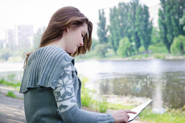 Fototapeta premium Girl sitting outdoors using a touchscreen tablet along a riverba
