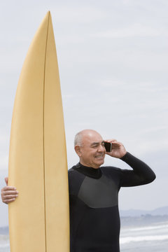Senior Hispanic man holding surfboard on beach and talking on cell phone