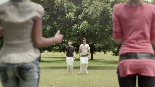 Young men playing sack race in park with girlfriends cheering