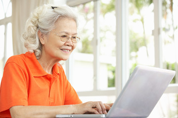 Senior Chinese Woman Sitting At Desk Using Laptop At Home