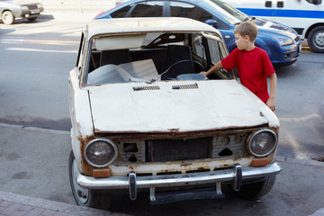 Boy in red T-shirt examines rusty with broken windshield car