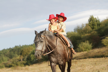 two happy children in cowboy hats riding horse on natural backgr