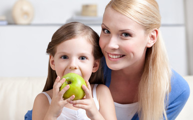 Smiley mom embraces her eating green apple daughter