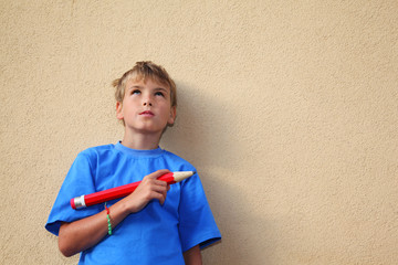 Boy with big red pencil stands near yellow wall and looks up.