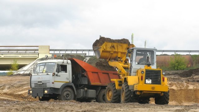 Dredger puts earth in dump truck in front of bridge
