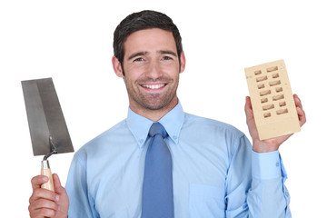 A man in a suit holding a brick and a trowel.