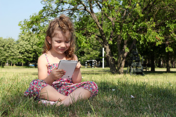 Fototapeta premium little girl sitting on grass and play with tablet pc