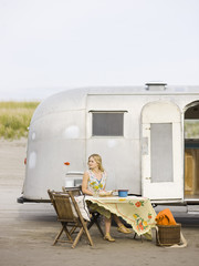 Naklejka premium woman sitting at a table on the beach outside her airstream classic trailer
