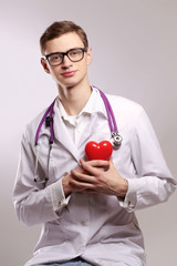 Male doctor with stethoscope holding heart on grey background.