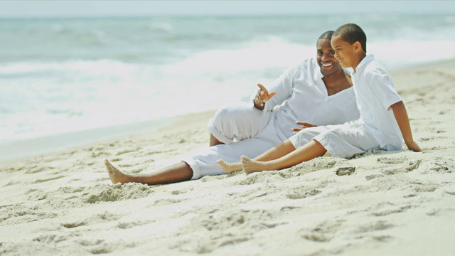 Ethnic son and his loving father sitting and laughing on beach