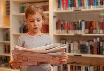 Little girl stands against bookshelves read newspaper