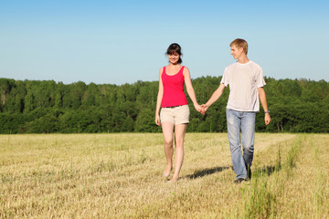 Husband, wife holding hands walk in field near wood