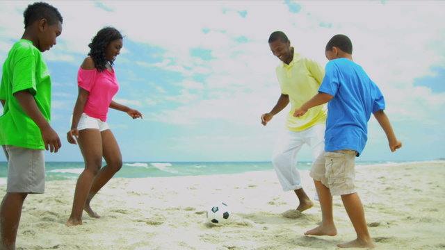 African American children enjoying summer kicking football with father on beach 
