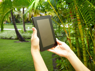 Hands with tablet computer in tropical garden.