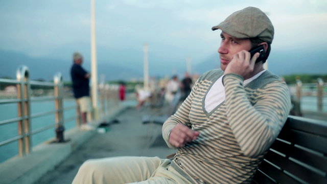 Happy young man sitting on bench and talking on cellphone, outdo