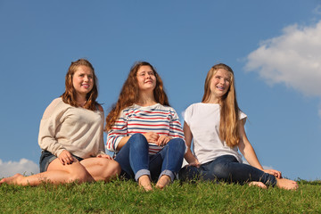 Three barefoot girls sit at green grass and look into distance