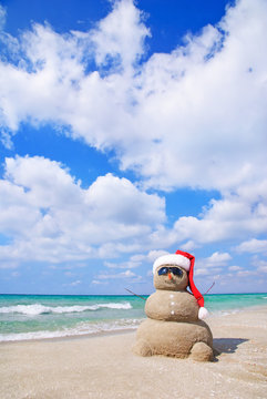 Smiling sandy snowman in red santa hat on the sea beach.