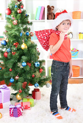 Little boy in Santa hat stands near Christmas tree holding bag