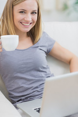 Fototapeta premium Smiling woman with coffee cup using laptop on sofa