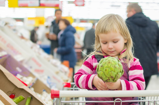 Adorable girl with artichoke in shopping cart in supermarket