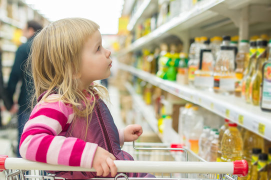 Adorable girl in shopping cart selecting sunflower oil in superm