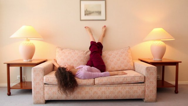 Young woman lies on sofa and shake legs at room with lamps on