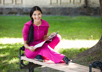 Cheerful young girl sitting on bench in park, reading magazine