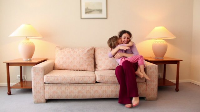 Little girl runs to her mother sit on sofa at room with lamp on