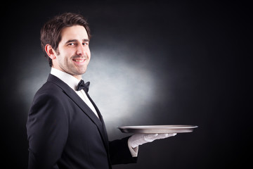 Portrait of a young waiter holding an empty dish on black backgr