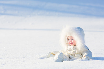 Obraz na płótnie Canvas Baby in white suit sitting in snow field on sunny winter day