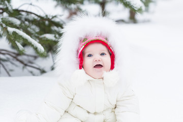 Sweet baby girl in white jacket sitting under snowy tree