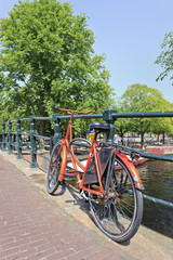 Fototapeta premium Orange bicycle parked against an iron railing in the ancient canal belt of Amsterdam, The Netherlands. 