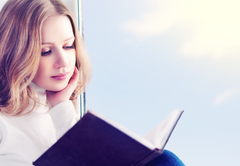 beautiful young woman reading a book while sitting at a window