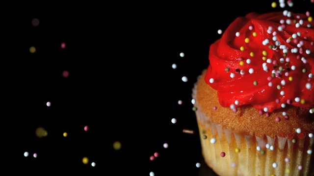 Colourful sprinkles pouring onto cupcake on black surface