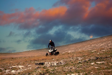 Man driving ATV in the meadows, red sunset clouds above, dramatic sunset, extreme motorsports background 