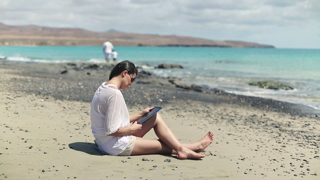 Woman with tablet computer sitting on the beach, family in the b