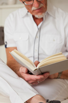 Senior man with glasses reading book in living room.