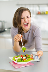 Laughing young woman eating salad and watching tv in kitchen