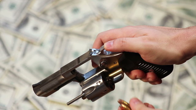 High angle of hands loading bullets into a handgun, over a background of hundred dollar bills.
