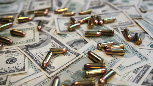 Gun bullets falling onto a pile of hundred dollar bills.