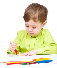 Little boy is drawing on white paper using crayon