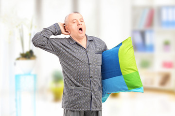Sleepy mature man in pajamas holding a pillow and yawning at hom
